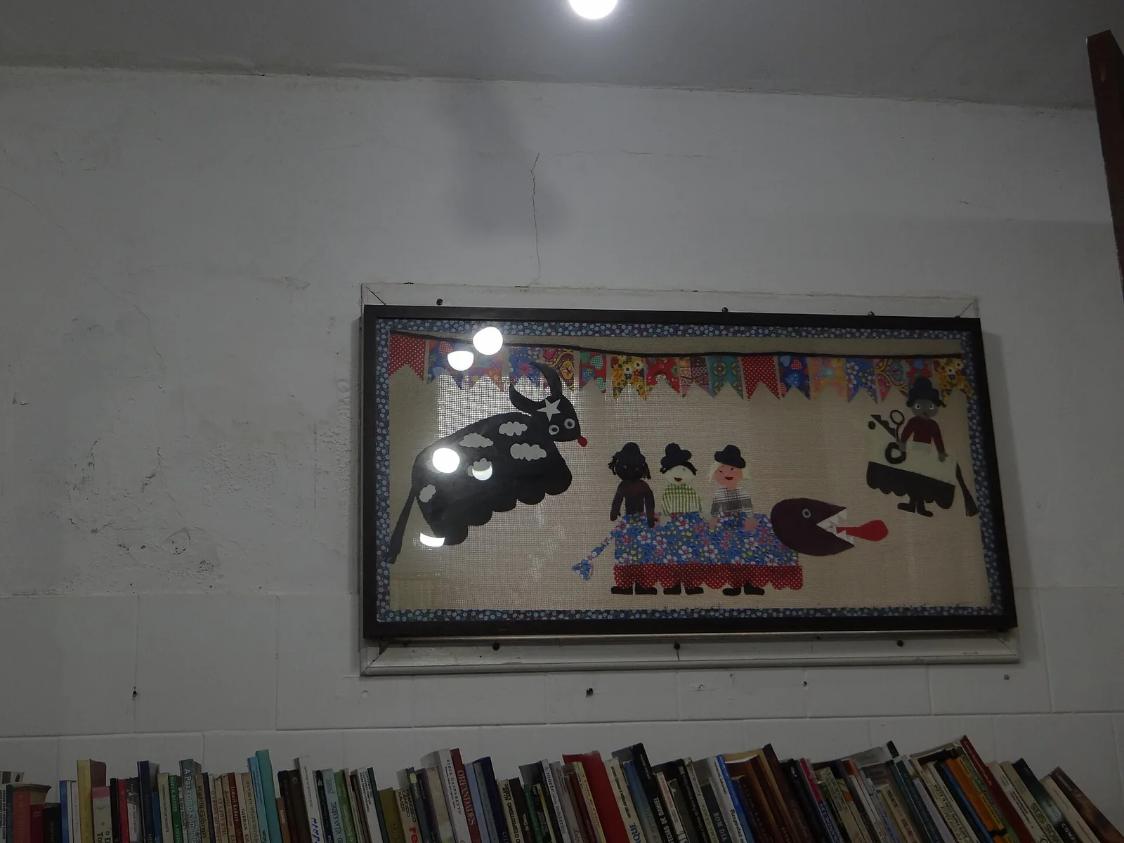 Painel de tecido emoldurado com personagens do boi de mamão, como o boi e a bernúncia, acima de uma estante de livros na Casa de Cultura de Garopaba