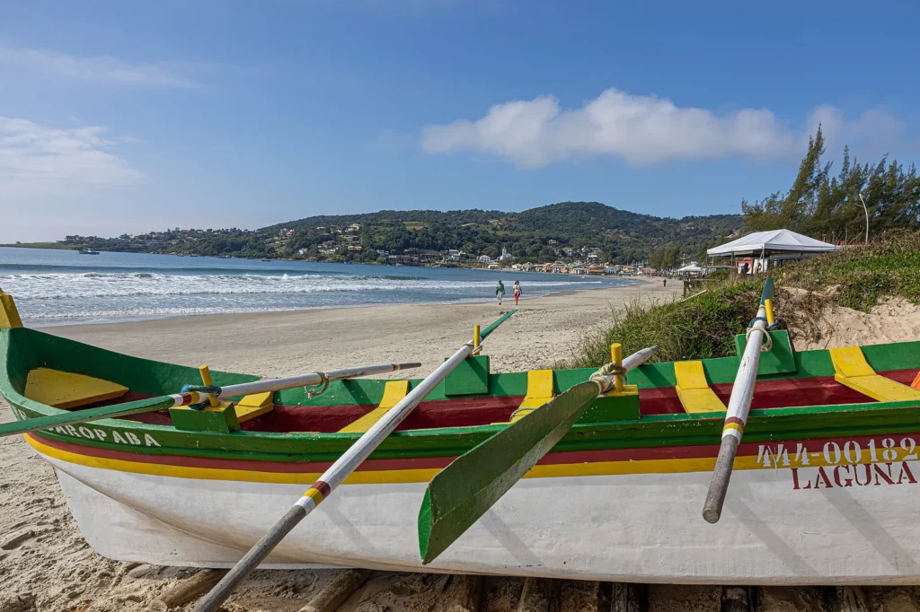 Canoa de pesca verde, amarela e vermelha com remos apoiada na areia da Praia de Garopaba; ao fundo, o mar, pessoas caminhando e o morro com casas
