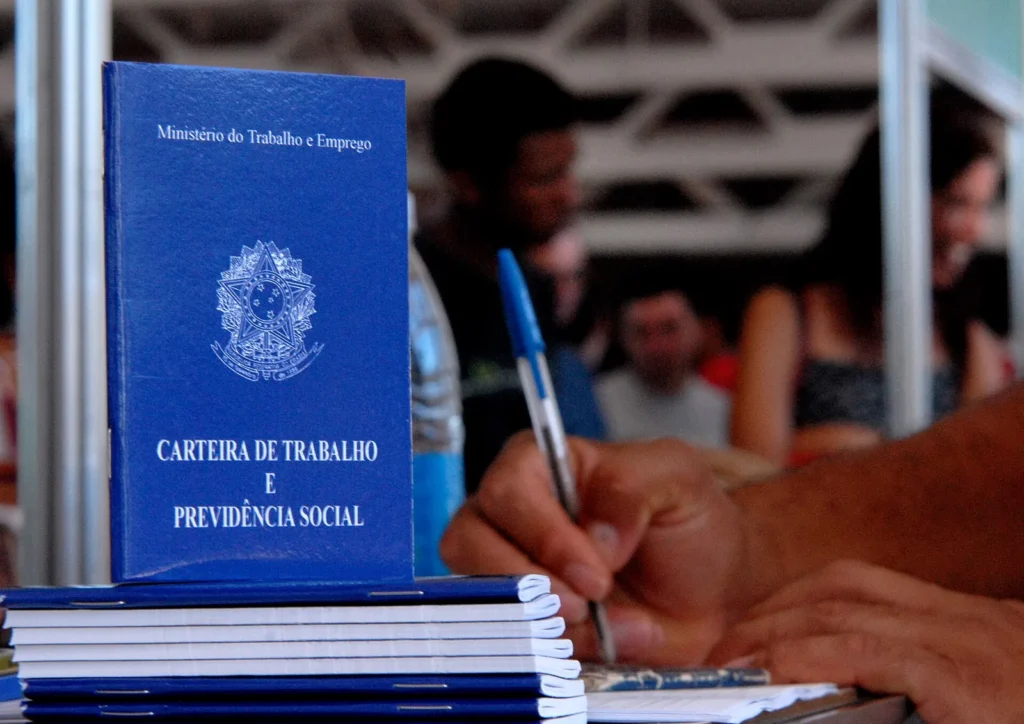 Pilha de carteiras de trabalho azuis sobre uma mesa enquanto uma pessoa preenche um documento