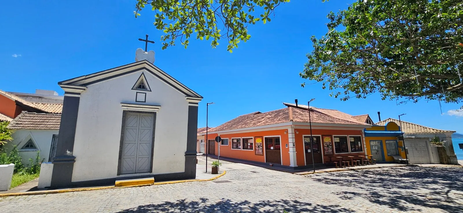 Fachada branca com pilastras cinza da Capela Senhor Bom Jesus, com porta dupla de madeira e cruz no alto, na rua de paralelepípedos do Centro Histórico de Garopaba