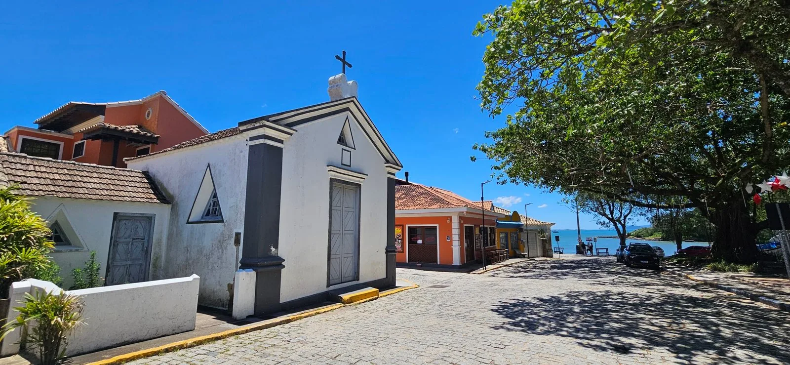 Capela Senhor Bom Jesus vista de lado, na rua de paralelepípedos sombreada por uma grande árvore, com casas coloridas e o mar da enseada de Garopaba ao fundo
