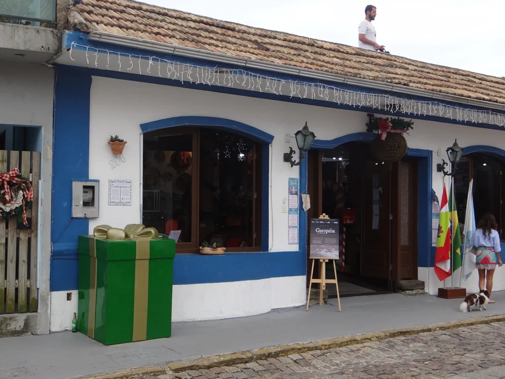 Fachada branca com barras azuis da Casa de Cultura de Garopaba, com janelas e portas em arco, lixeiras verdes e calçada de pedra