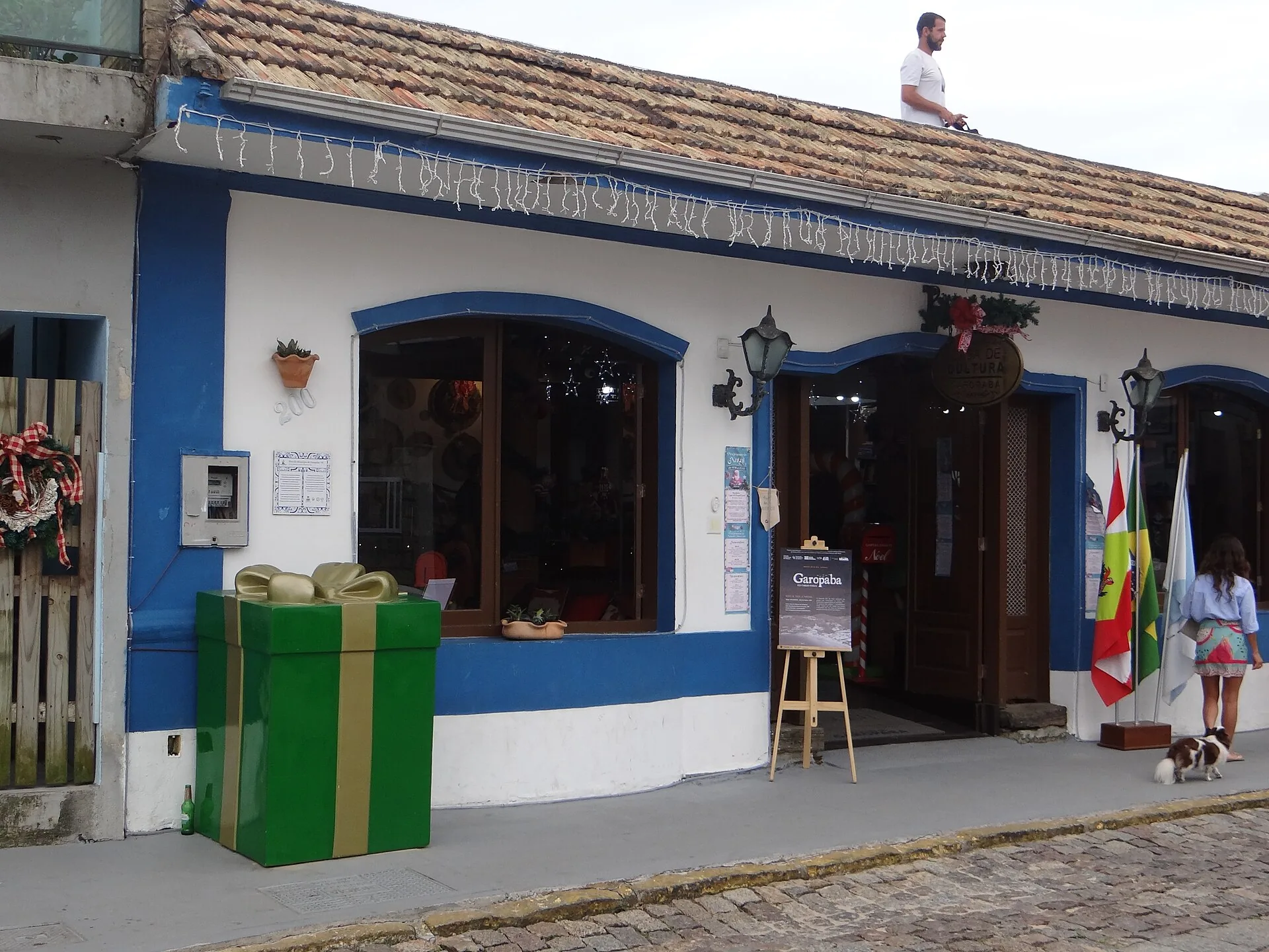 Fachada branca com barras azuis da Casa de Cultura de Garopaba, com janelas e portas em arco, lixeiras verdes e calçada de pedra