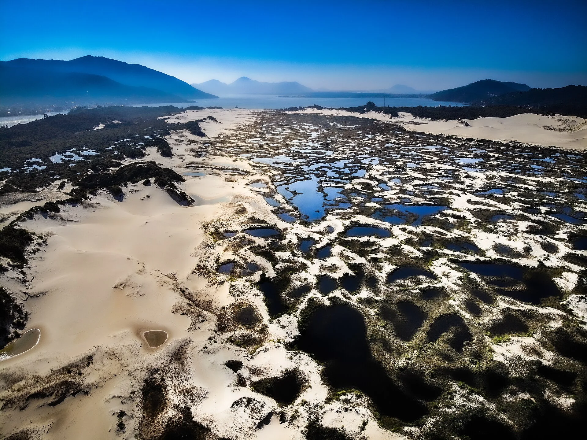 Vista aérea do campo de dunas com poças e lagoas temporárias entre a areia, e morros ao fundo