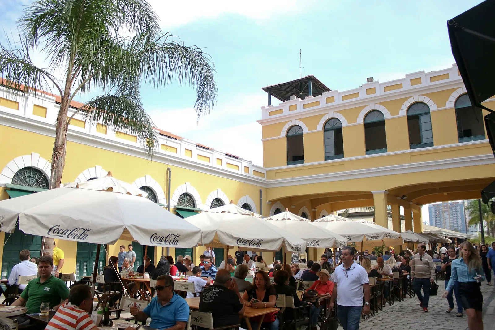 Vão central do Mercado Público de Florianópolis com mesas, guarda-sóis e pessoas almoçando junto ao casarão amarelo