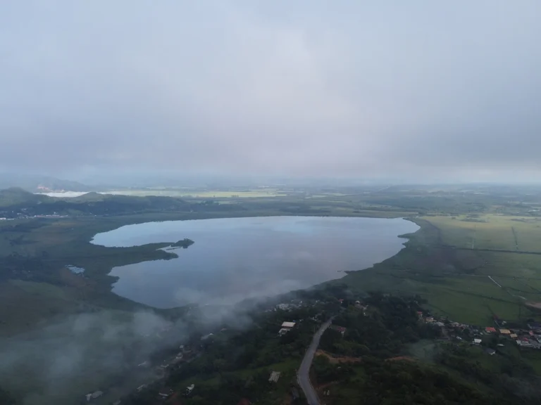 Vista aérea da Lagoa do Ribeirão cercada de campos e mata, com névoa baixa sobre os morros e a estrada em primeiro plano