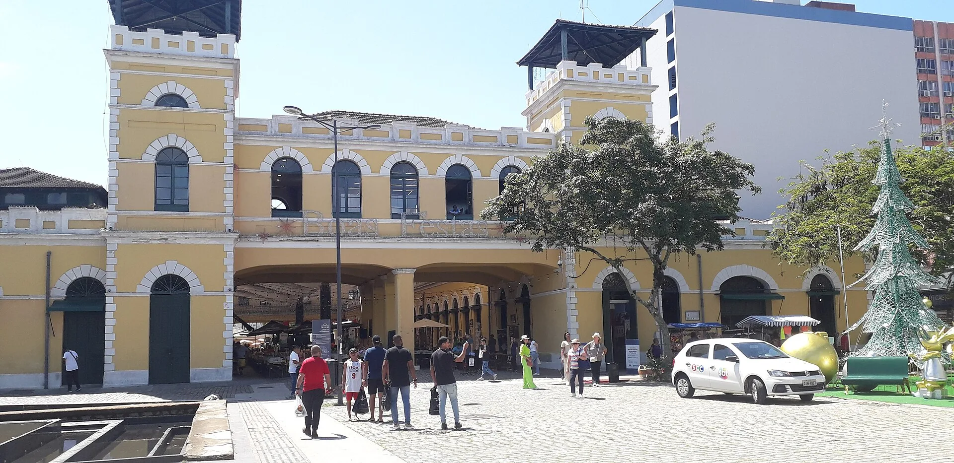 Fachada amarela do Mercado Público de Florianópolis com torre central, árvores e pessoas no calçadão em frente