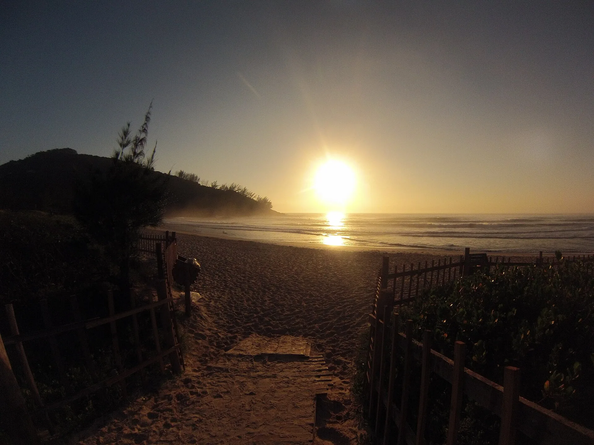 Nascer do sol sobre o mar visto de um caminho de areia com cercas de madeira que leva à Praia da Ferrugem