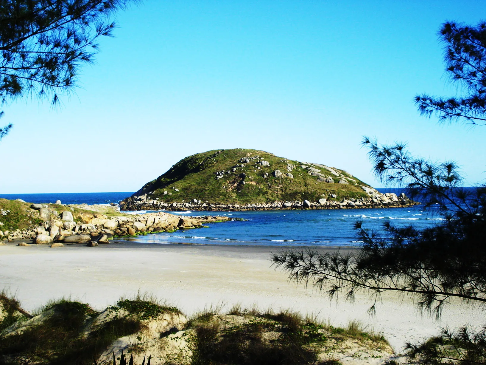 Praia da Vila com mar azul, pedras no canto e uma ilha coberta de vegetação próxima à costa, sob céu azul