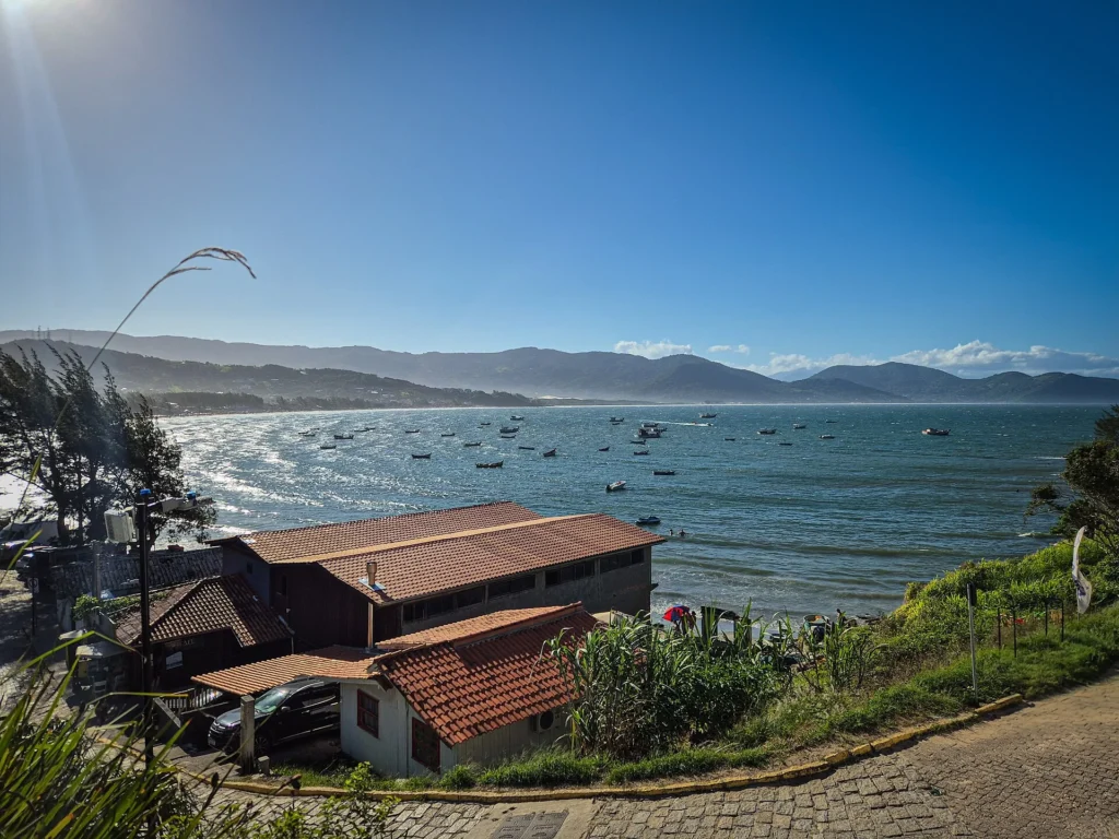 Vista do alto da enseada de Garopaba, com barcos ancorados no mar calmo, casas de telhado laranja e a rua da orla