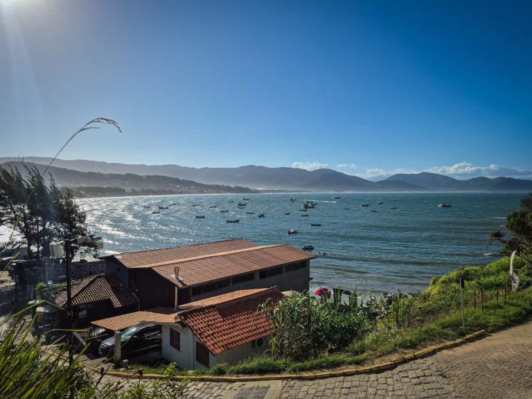 Vista do alto da enseada de Garopaba, com barcos ancorados no mar calmo, casas de telhado laranja e a rua da orla